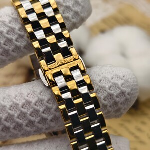 Puede incluir: Primer plano de una correa de reloj dorada y plateada con el nombre de la marca "VERSACE" visible. La correa presenta un patr&oacute;n repetitivo de eslabones rectangulares dorados y plateados, creando una apariencia texturizada. La correa del reloj est&aacute; sujeta por una tela blanca.