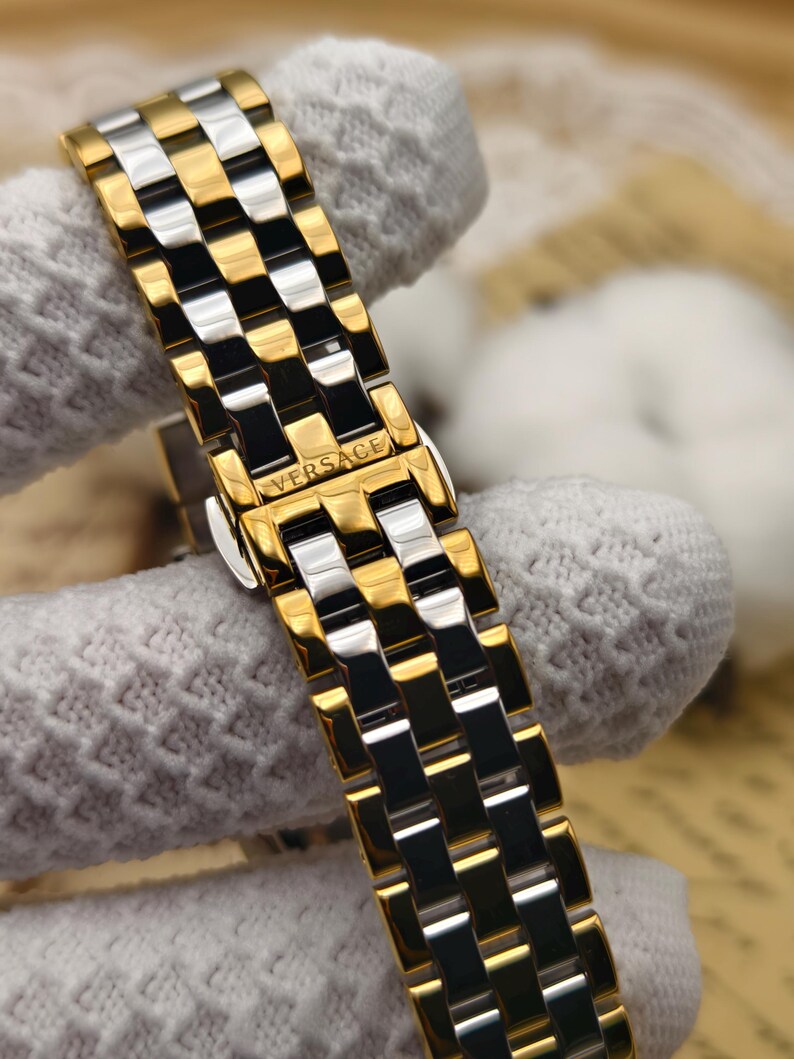 Puede incluir: Primer plano de una correa de reloj dorada y plateada con el nombre de la marca "VERSACE" visible. La correa presenta un patr&oacute;n repetitivo de eslabones rectangulares dorados y plateados, creando una apariencia texturizada. La correa del reloj est&aacute; sujeta por una tela blanca.