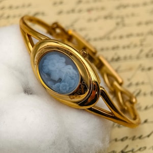 May include: A gold-tone bangle watch featuring an oval face with a light blue cameo inlay depicting a profile portrait. The watch has a polished metal finish, a slender band, and the word Cameo is printed in gold on the lower right side of the bezel.