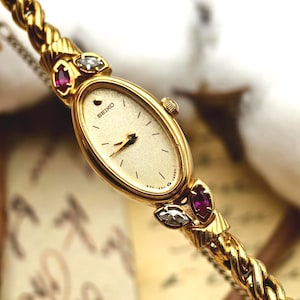 May include: A gold-tone Seiko watch with an oval face, a textured cream dial, and gold hands. The watch features a twisted chain-link band accented with marquise-cut red and clear stones at the lugs. The dial displays the Seiko logo and Japan text.