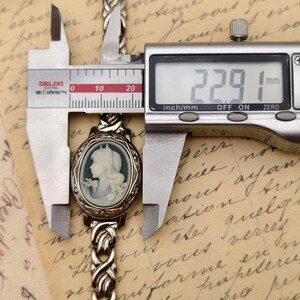 Puede incluir: Una pulsera de metal plateado con un colgante de camafeo ovalado que muestra el retrato de perfil de una figura. Un calibrador digital est&aacute; colocado sobre el colgante, mostrando una medida de 22,91 mm en su pantalla, indicando su anchura.