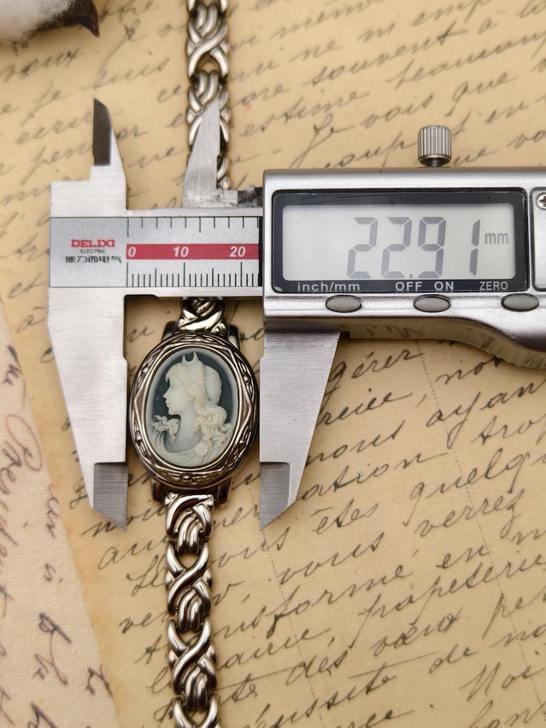 Puede incluir: Una pulsera de metal plateado con un colgante de camafeo ovalado que muestra el retrato de perfil de una figura. Un calibrador digital est&aacute; colocado sobre el colgante, mostrando una medida de 22,91 mm en su pantalla, indicando su anchura.