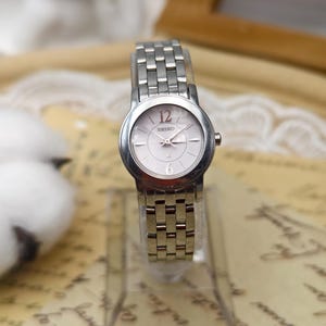 May include: A silver Seiko wristwatch with a white face, black hands, and red numerals at 12 and 6 o'clock. The watch has a rectangular link band and is displayed on a clear stand. The watch face has the word "SEIKO" printed on it.