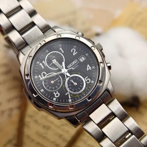 Op de afbeelding: Een Seiko chronograaf horloge van roestvrij staal met een zwarte wijzerplaat. Het horloge heeft meerdere sub-wijzerplaten, lichtgevende wijzers en een datumweergave. De band en de lunette zijn van roestvrij staal.