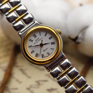 May include: A silver and gold-toned wristwatch with a white face and Roman numeral hour markers. The watch features the text "YOU Sporty Elegance" and "Quartz". The watch band is a combination of silver and gold links.