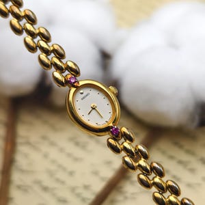 May include: A gold-tone Seiko wristwatch with an oval white dial, gold hands, and hour markers. The watch features a link bracelet and two small pink stones set at the top and bottom of the oval case where the bracelet connects to the watch head.