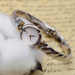 May include: A Tisse wristwatch featuring a round white dial with gold hands and a Roman numeral at the twelve o'clock position. The watch has a two-tone silver and gold twisted metal band with small dark purple accents at the lugs.