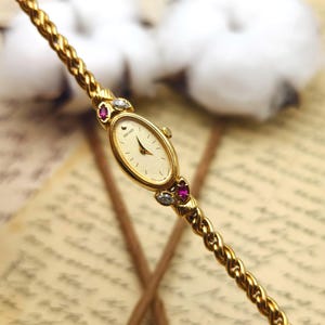 May include: A gold-tone Seiko wristwatch with an oval face and a textured cream dial. The watch features a twisted rope-style chain band accented with marquise-cut red and clear stones at the lugs. The watch hands and hour markers are gold.