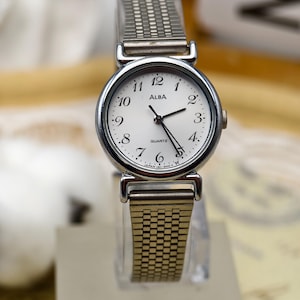 May include: A silver ALBA quartz watch with a white face and black numerals. The watch has a stretch band and is displayed on a clear stand. The watch face reads "ALBA QUARTZ".