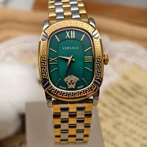 Puede incluir: Un reloj Versace dorado y plateado con una esfera verde rectangular. El reloj presenta n&uacute;meros romanos dorados, manecillas doradas y el logotipo de la cabeza de Medusa de Versace. La correa del reloj es una mezcla de eslabones dorados y plateados.