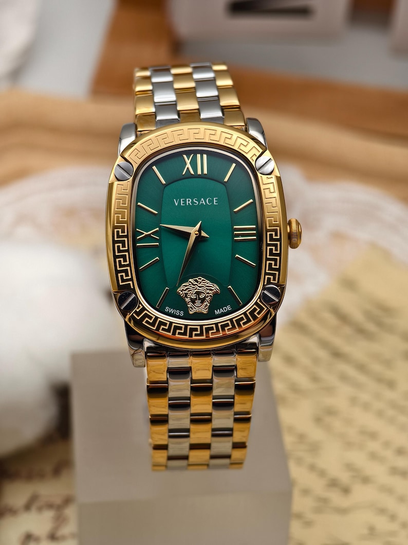 Puede incluir: Un reloj Versace dorado y plateado con una esfera verde rectangular. El reloj presenta n&uacute;meros romanos dorados, manecillas doradas y el logotipo de la cabeza de Medusa de Versace. La correa del reloj es una mezcla de eslabones dorados y plateados.