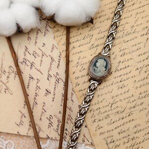 Puede incluir: Un reloj de pulsera de tono plateado con una esfera ovalada que presenta un retrato de perfil en camafeo azul y blanco. La correa del reloj est&aacute; construida con eslabones met&aacute;licos plateados interconectados con un patr&oacute;n trenzado.