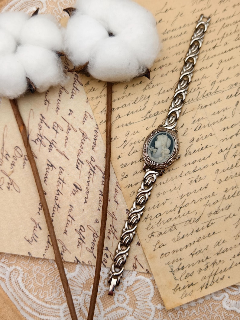 Puede incluir: Un reloj de pulsera de tono plateado con una esfera ovalada que presenta un retrato de perfil en camafeo azul y blanco. La correa del reloj est&aacute; construida con eslabones met&aacute;licos plateados interconectados con un patr&oacute;n trenzado.