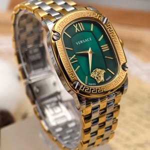 Puede incluir: Un reloj Versace bicolor dorado y plateado con una esfera verde rectangular. El reloj presenta n&uacute;meros romanos dorados, un emblema de cabeza de Medusa dorada y la palabra "Versace" en blanco. La correa del reloj es una combinaci&oacute;n de eslabones dorados y plateados.