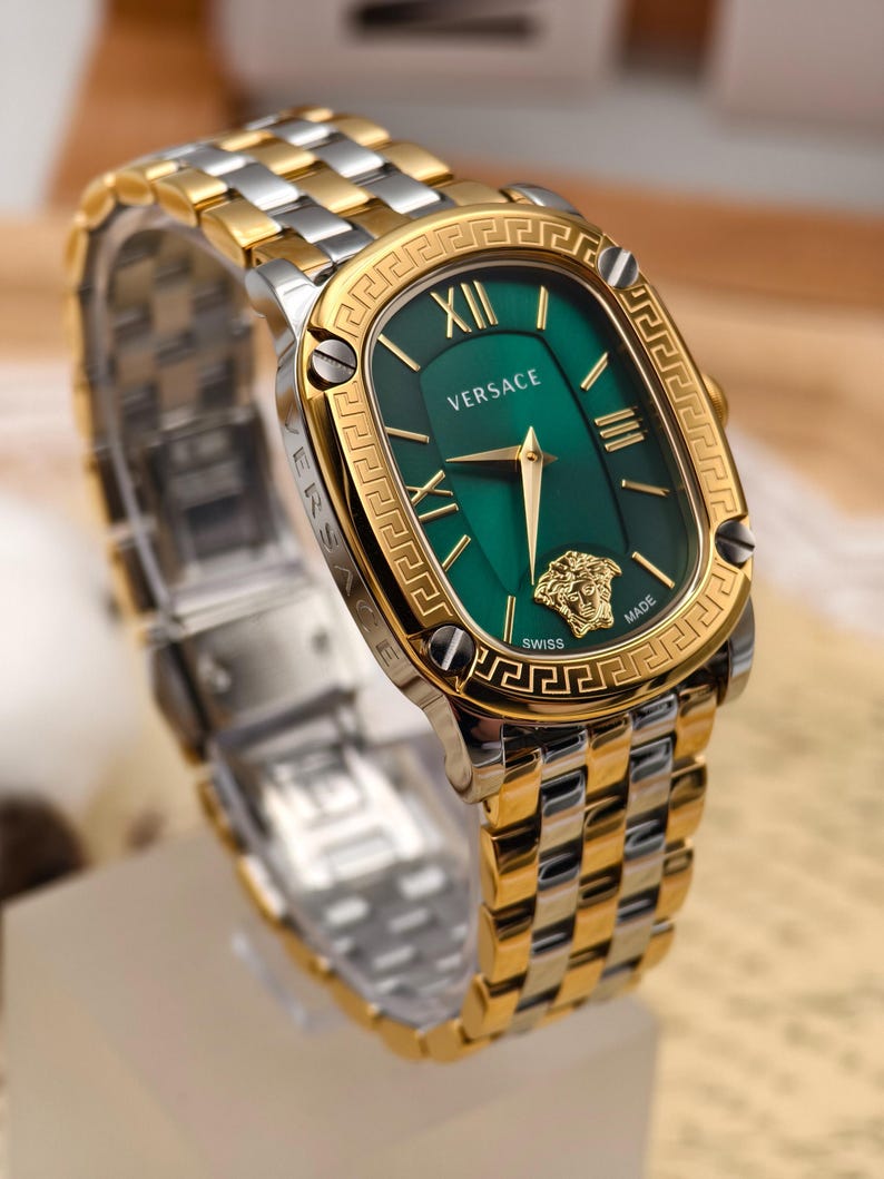 Puede incluir: Un reloj Versace bicolor dorado y plateado con una esfera verde rectangular. El reloj presenta n&uacute;meros romanos dorados, un emblema de cabeza de Medusa dorada y la palabra "Versace" en blanco. La correa del reloj es una combinaci&oacute;n de eslabones dorados y plateados.