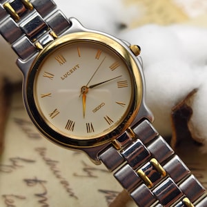 Puede incluir: Un reloj de pulsera Seiko Lucent con una correa de metal plateado y dorado. La esfera crema presenta números romanos y agujas doradas. La correa está hecha de eslabones rectangulares con detalles dorados.
