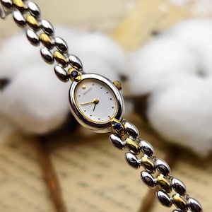 May include: A Seiko wristwatch with an oval white dial, gold-tone hands, and a two-tone silver and gold link bracelet. The watch case features small blue accents at the top and bottom lugs, with a gold-tone crown on the right side of the case.