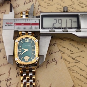 Puede incluir: Un reloj Versace dorado y plateado con esfera verde, n&uacute;meros romanos y el logotipo de Versace. El reloj se mide con un calibrador digital, mostrando un ancho de 29,17 mm.