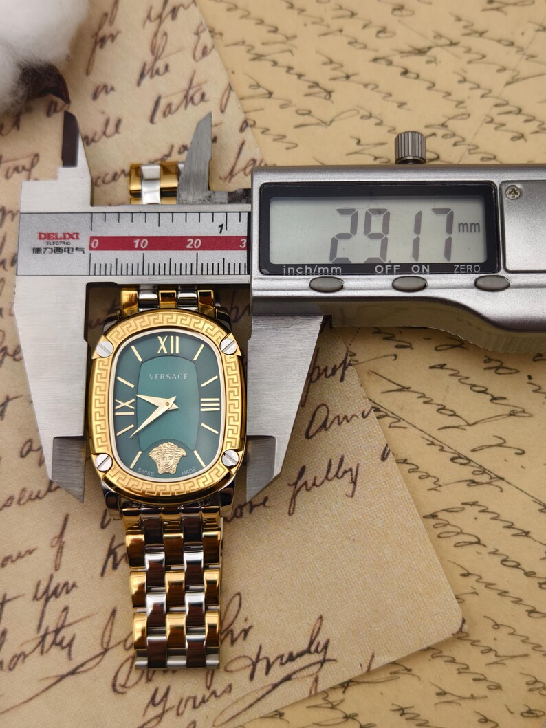 Puede incluir: Un reloj Versace dorado y plateado con esfera verde, n&uacute;meros romanos y el logotipo de Versace. El reloj se mide con un calibrador digital, mostrando un ancho de 29,17 mm.