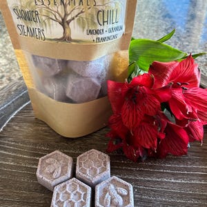 May include: A bag of purple shower steamers with the text "Willow Roots Essentials Shower Steamers Chill Lavender + Geranium + Frankincense" on the front. Four purple shower steamers are arranged in a honeycomb pattern on a wooden surface.