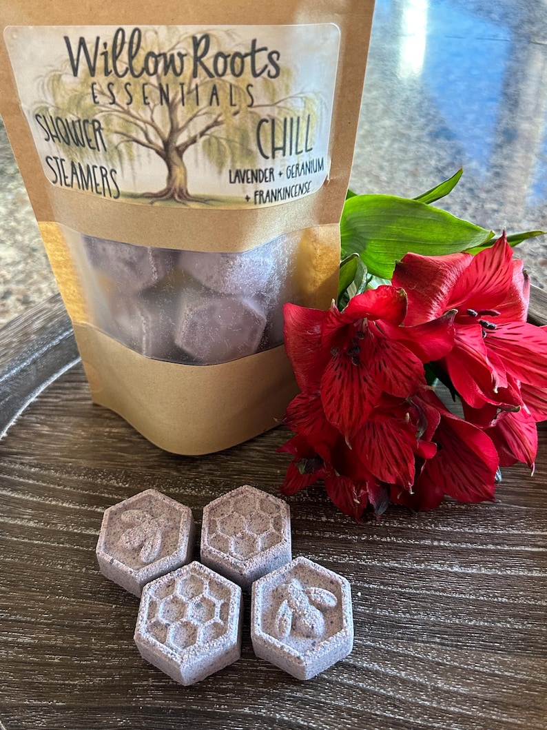 May include: A bag of purple shower steamers with the text "Willow Roots Essentials Shower Steamers Chill Lavender + Geranium + Frankincense" on the front. Four purple shower steamers are arranged in a honeycomb pattern on a wooden surface.