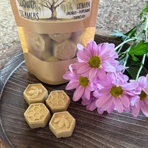 May include: Four hexagonal shower steamers with a bee design on two of them. The steamers are on a wooden surface with a bag of shower steamers in the background. The bag is labeled "Willow Roots Essentials - Migraine Hitman Lavender + Peppermint + Ravintsara"