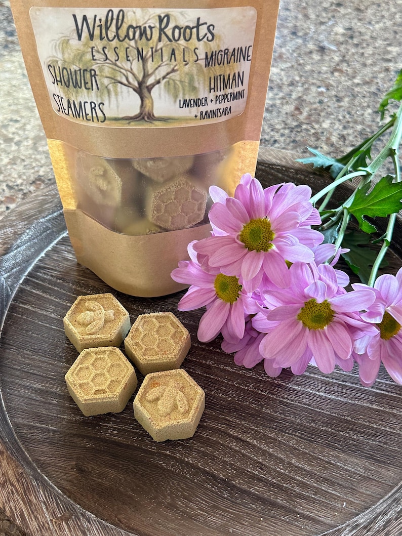 May include: Four hexagonal shower steamers with a bee design on two of them. The steamers are on a wooden surface with a bag of shower steamers in the background. The bag is labeled "Willow Roots Essentials - Migraine Hitman Lavender + Peppermint + Ravintsara"