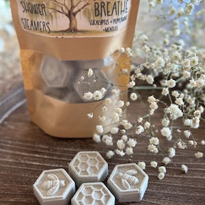 May include: A bag of shower steamers with a label that reads "Willow Roots Essentials Breathe Eucalyptus + Peppermint + Menthol Shower Steamers". The steamers are hexagonal and have a bee design. The bag is sitting on a wooden surface with white flowers.