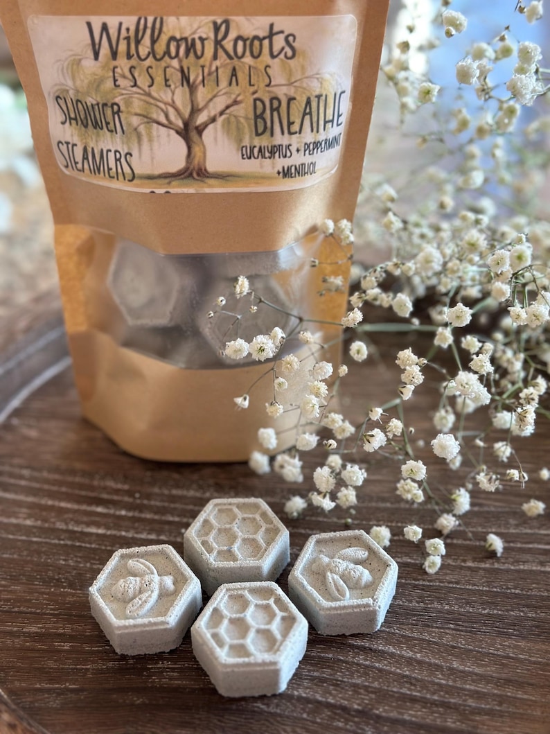 May include: A bag of shower steamers with a label that reads "Willow Roots Essentials Breathe Eucalyptus + Peppermint + Menthol Shower Steamers". The steamers are hexagonal and have a bee design. The bag is sitting on a wooden surface with white flowers.