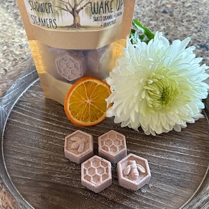 May include: Four purple shower steamers shaped like honeycombs with a bee design. The steamers are on a wooden tray with a sliced orange and a white flower.