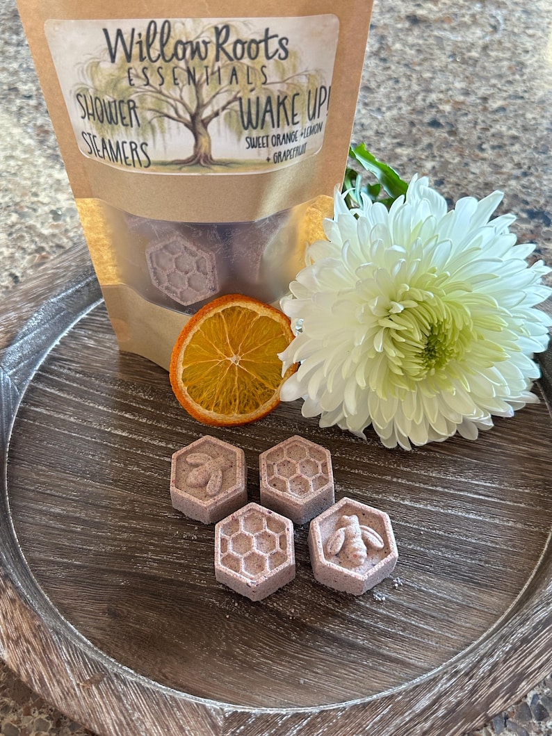 May include: Four purple shower steamers shaped like honeycombs with a bee design. The steamers are on a wooden tray with a sliced orange and a white flower.