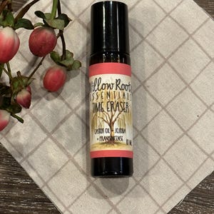 May include: A small, clear glass bottle with a black rollerball top. The bottle is labeled "Willow Root Essentials Time Eraser" and lists the ingredients as "Castor Oil, Jojoba, Frankincense". The bottle is sitting on a light brown fabric with a checkered pattern.