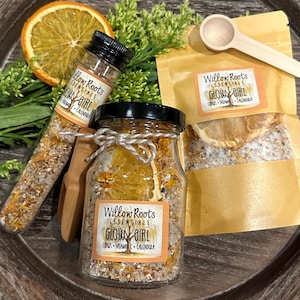 May include: Two glass jars filled with bath salts and dried citrus fruit. The jars have labels that read "Willow Roots Essentials Glow Girl Citrus Vitamin C Calendula".