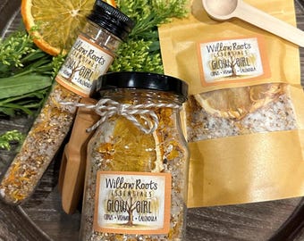 May include: Two glass jars filled with bath salts and dried citrus fruit. The jars have labels that read "Willow Roots Essentials Glow Girl Citrus Vitamin C Calendula".