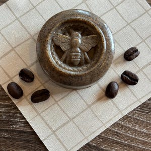 May include: A round brown soap bar with a bee design in the center. The soap is surrounded by coffee beans on a beige and brown checkered surface.