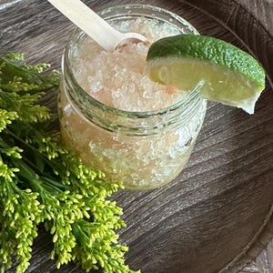 May include: A glass jar filled with a pink and white slush, topped with a lime wedge and a wooden spoon. The jar is sitting on a wooden tray with green foliage.