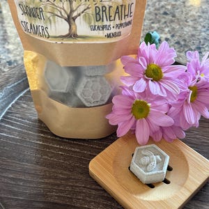 May include: A bag of shower steamers labeled "Willow Roots Essentials" with the scent "Breathe Eucalyptus + Peppermint + Menthol". The bag contains six gray hexagonal shower steamers with a bee design. The bag is sitting on a wooden surface with a single shower steamer on a small wooden tray.