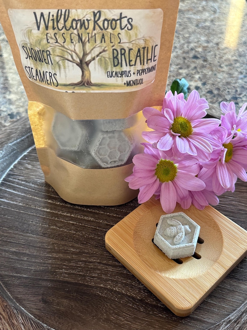 May include: A bag of shower steamers labeled "Willow Roots Essentials" with the scent "Breathe Eucalyptus + Peppermint + Menthol". The bag contains six gray hexagonal shower steamers with a bee design. The bag is sitting on a wooden surface with a single shower steamer on a small wooden tray.