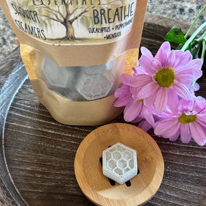 May include: A brown paper bag with a white label that reads "Willow Roots Essentials Shower Steamers Breathe Eucalyptus + Peppermint + Menthol". The bag contains six white shower steamers shaped like hexagons. The bag is sitting on a wooden surface with a pink flower.