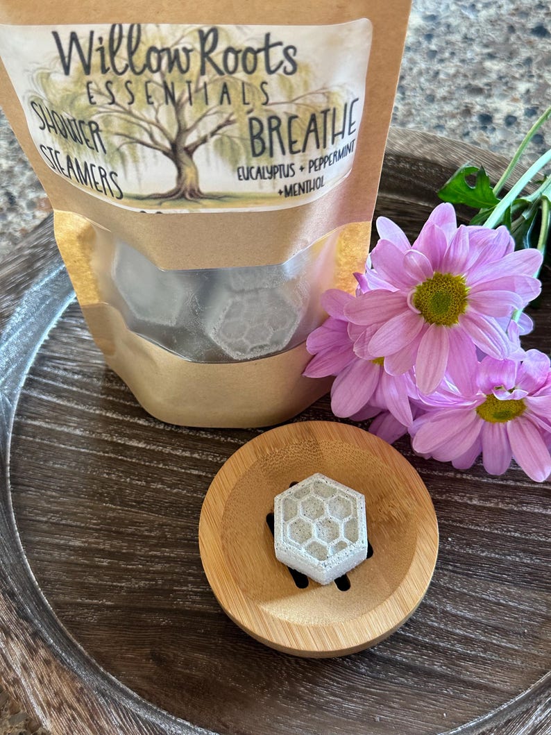 May include: A brown paper bag with a white label that reads "Willow Roots Essentials Shower Steamers Breathe Eucalyptus + Peppermint + Menthol". The bag contains six white shower steamers shaped like hexagons. The bag is sitting on a wooden surface with a pink flower.