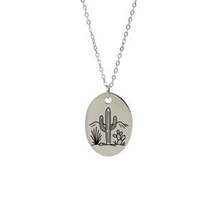 May include: Silver necklace with an oval pendant depicting a desert scene. The pendant has a black line drawing of a cactus, other desert plants, and mountains. The chain is a delicate silver link, suitable for everyday wear.