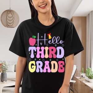 Peut inclure: Un t-shirt noir arborant un motif coloré avec le texte "Hello THIRD GRADE" dans une police arrondie et ludique. Le design comprend une pomme rose, un petit nœud rouge et une cloche jaune, agrémenté de petites étoiles et d'éclats multicolores.