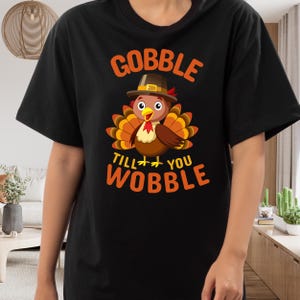 May include: A black short-sleeved t-shirt featuring a graphic of a brown turkey wearing a pilgrim hat. The text GOBBLE TILL YOU WOBBLE is printed in orange above and below the turkey illustration, which also includes yellow and red accents.
