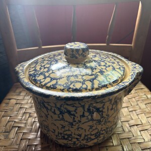 Puede incluir: Un tarro de cerámica azul y blanco con tapa. El tarro tiene un patrón moteado y está sentado sobre una superficie tejida.