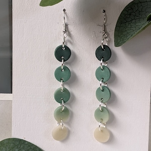 May include: A pair of green and white ombre dangle earrings with silver hoops. Each earring has five round clay beads that fade from dark green to white.