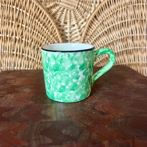 May include: A small ceramic mug with a green speckled pattern and a black rim. The handle and interior are white. The mug is set on a dark wooden surface, with a woven wicker background.
