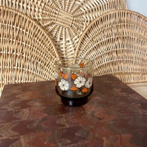 May include: A vintage-style glass tumbler with a floral pattern. The clear glass features white and orange flower designs. The tumbler sits on a dark brown base, with a woven wicker background.