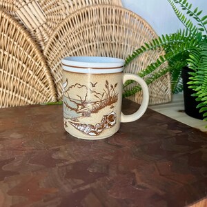 May include: A beige ceramic mug with a coastal design featuring seashells, birds, and a landscape scene. The mug has a white interior and a brown band near the rim. The handle is a matching beige color.