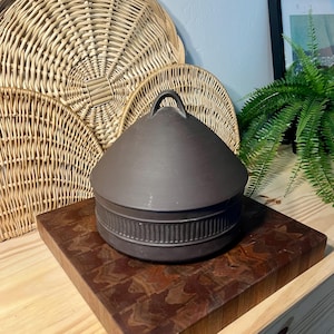 May include: A dark brown ceramic lidded pot with a handle on top. The pot has a ridged band around the base. Woven wicker baskets are in the background.