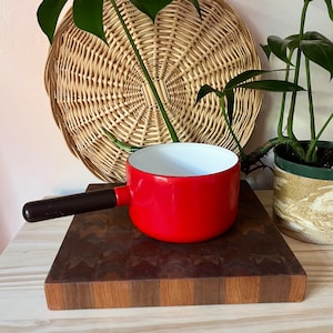 May include: A red enamel saucepan with a black handle. The saucepan is sitting on a wooden cutting board.